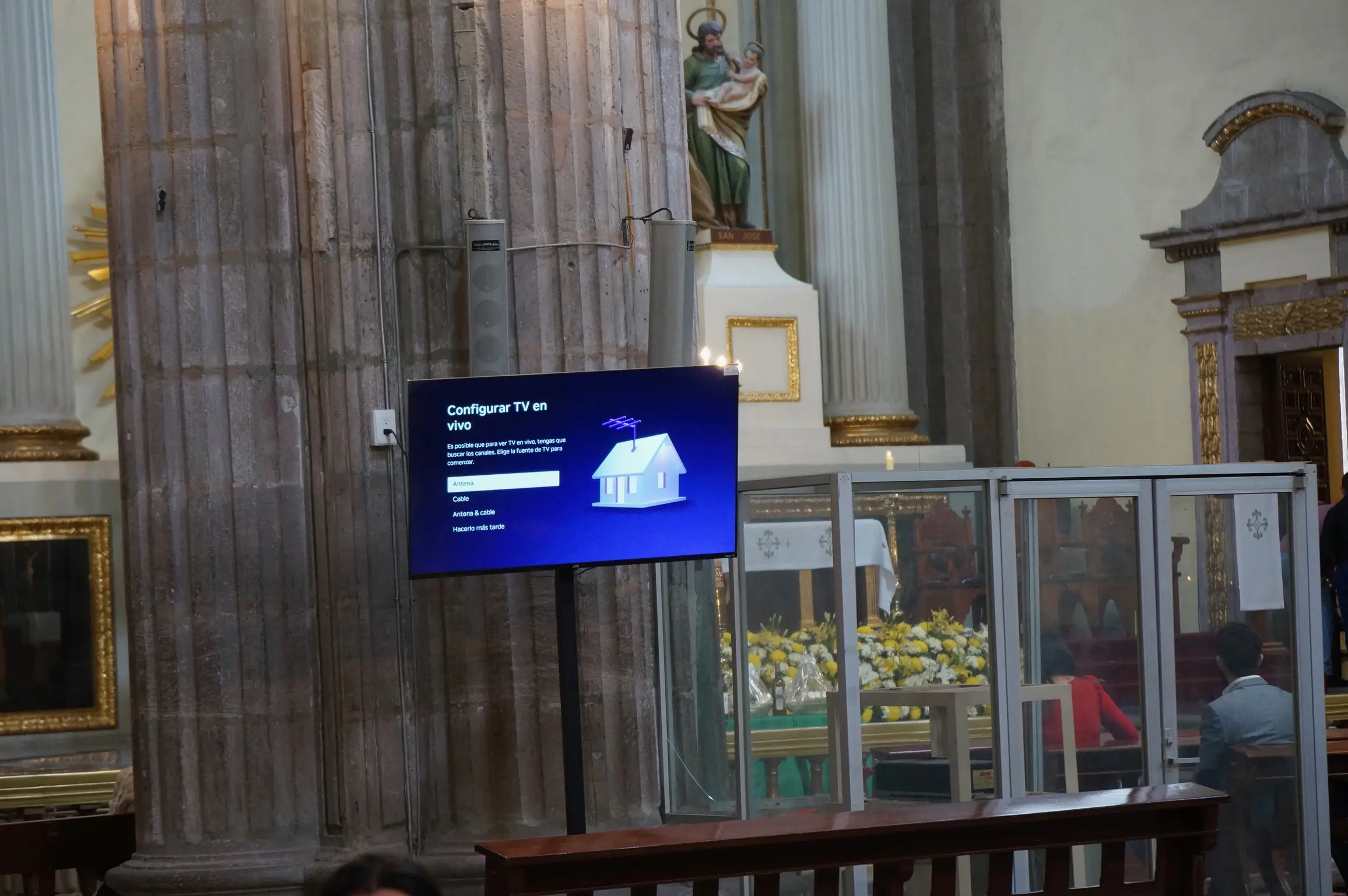 In the foreground, a TV with a purple screen says “Configurar TV en vivo: Es posible que para ver TV en vivo, tengas que buscar los canales. Elige la fuente de TV para comenzar. - Antena - Cable - Antena & cable - Hacerio más tarde”. Behind the TV is a white and gold statue of Jesus with other ornate and gold-plated decorations beside it.
