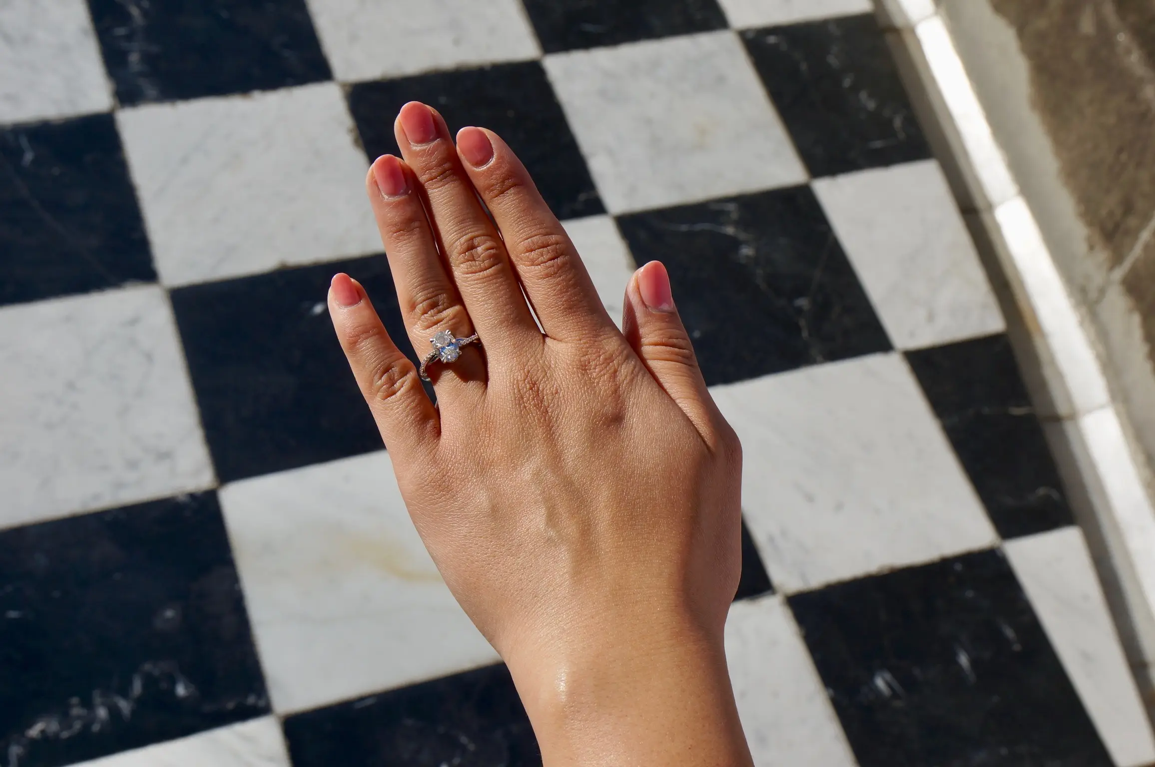 A woman’s hand with pink-painted nails hovers above a black and white checkered floor. On her ring finger is a diamond engagement ring.