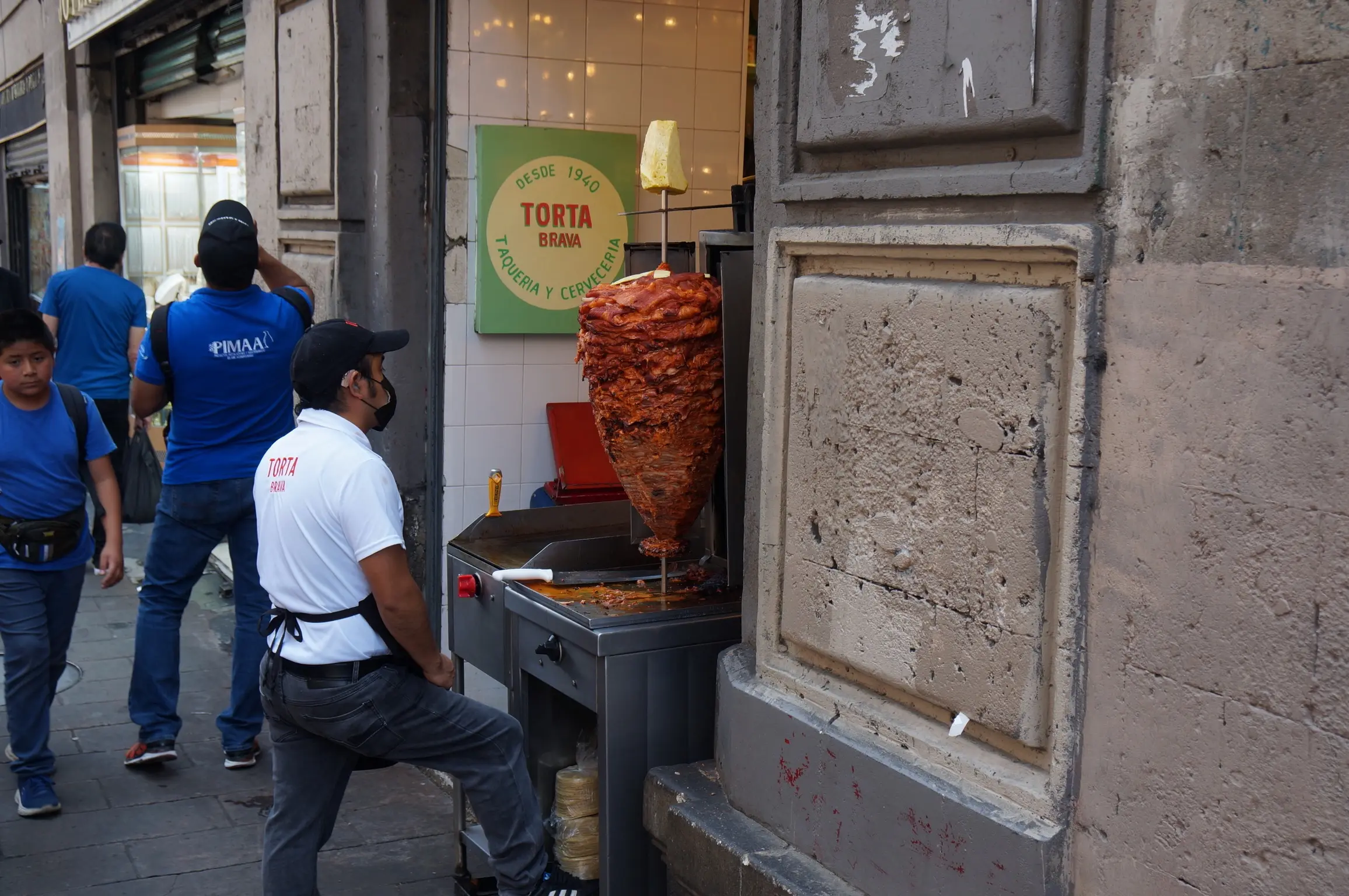 A man in a white shirt stands on the street next to a tropo of red meat, large at the top and thinner at the bottom, forming a cone shape. The sign on the side of the wall reads “Desde 1940. Torta Brava. Taqueria y Cerveceria. Several people in blue shirts walk on the sidewalk behind him.”