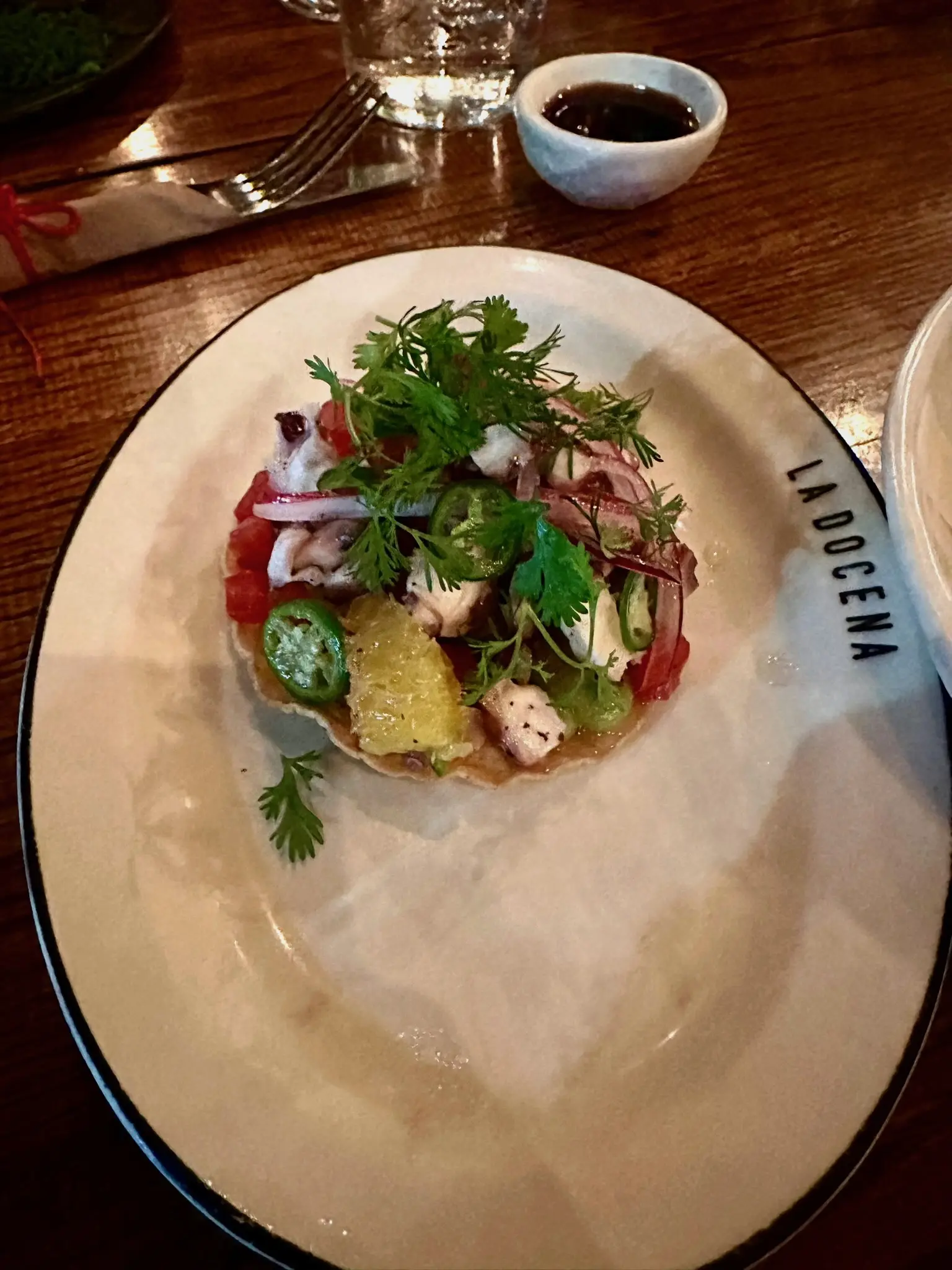 An ovular white plate with the text “LA DOCENA” on the rim sits under a tostada with pineapple, red onion slices, jalapeño slices, grilled octopus slices, and topped with cilantro.