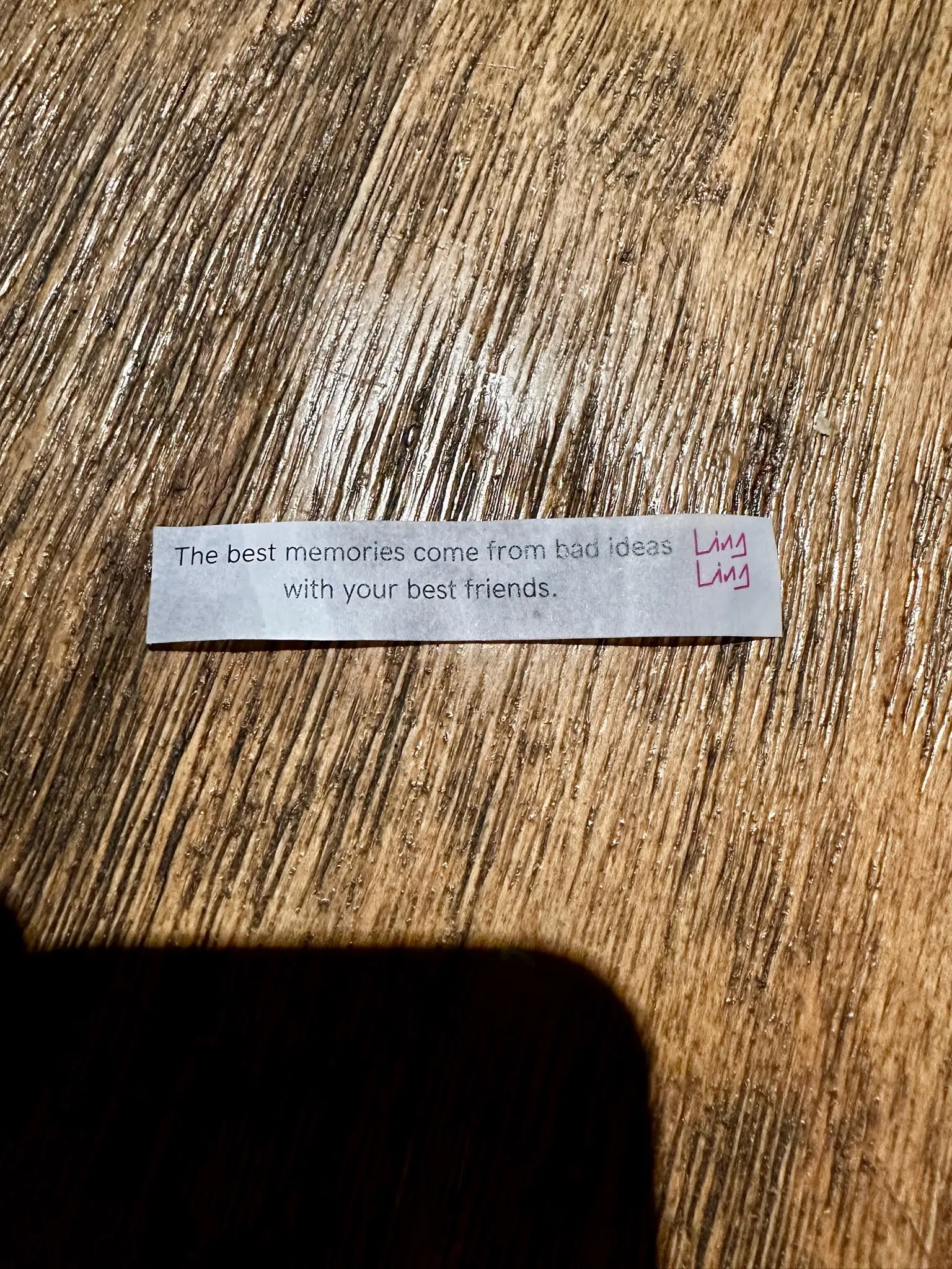 A strip of paper sits on a wooden table. The paper says “The best memories come from bad ideas with your best friends.” with the Ling Ling logo in the upper right corner.