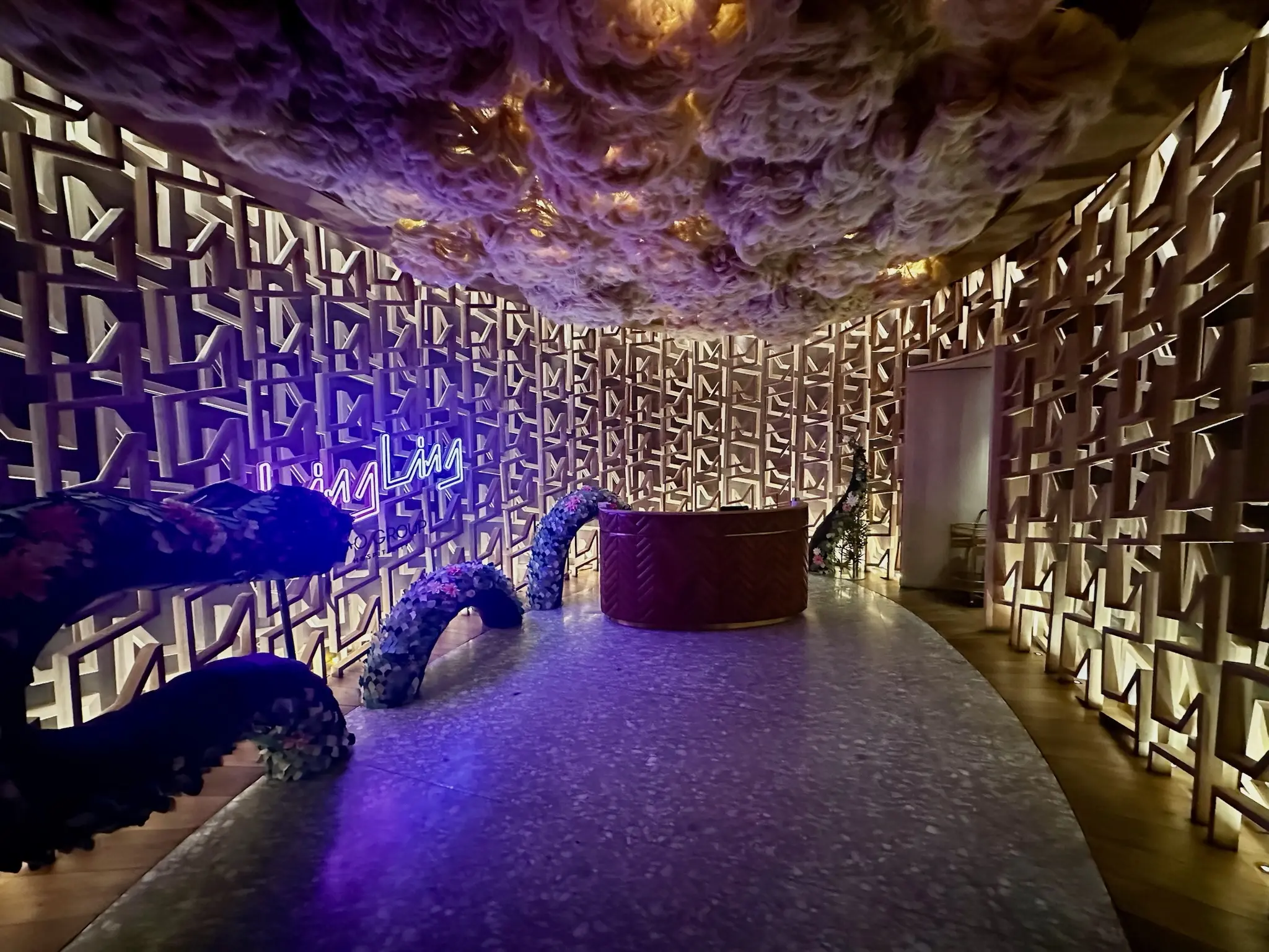 A dimly lit lobby with a fake glowing cloud ceiling and a patterned, textured wall with a blue neon sign that says “Ling Ling” on the wall on the left. Around the sign is a dragon made out of a topiary that looks like it’s dissapearing into and out of the floor.