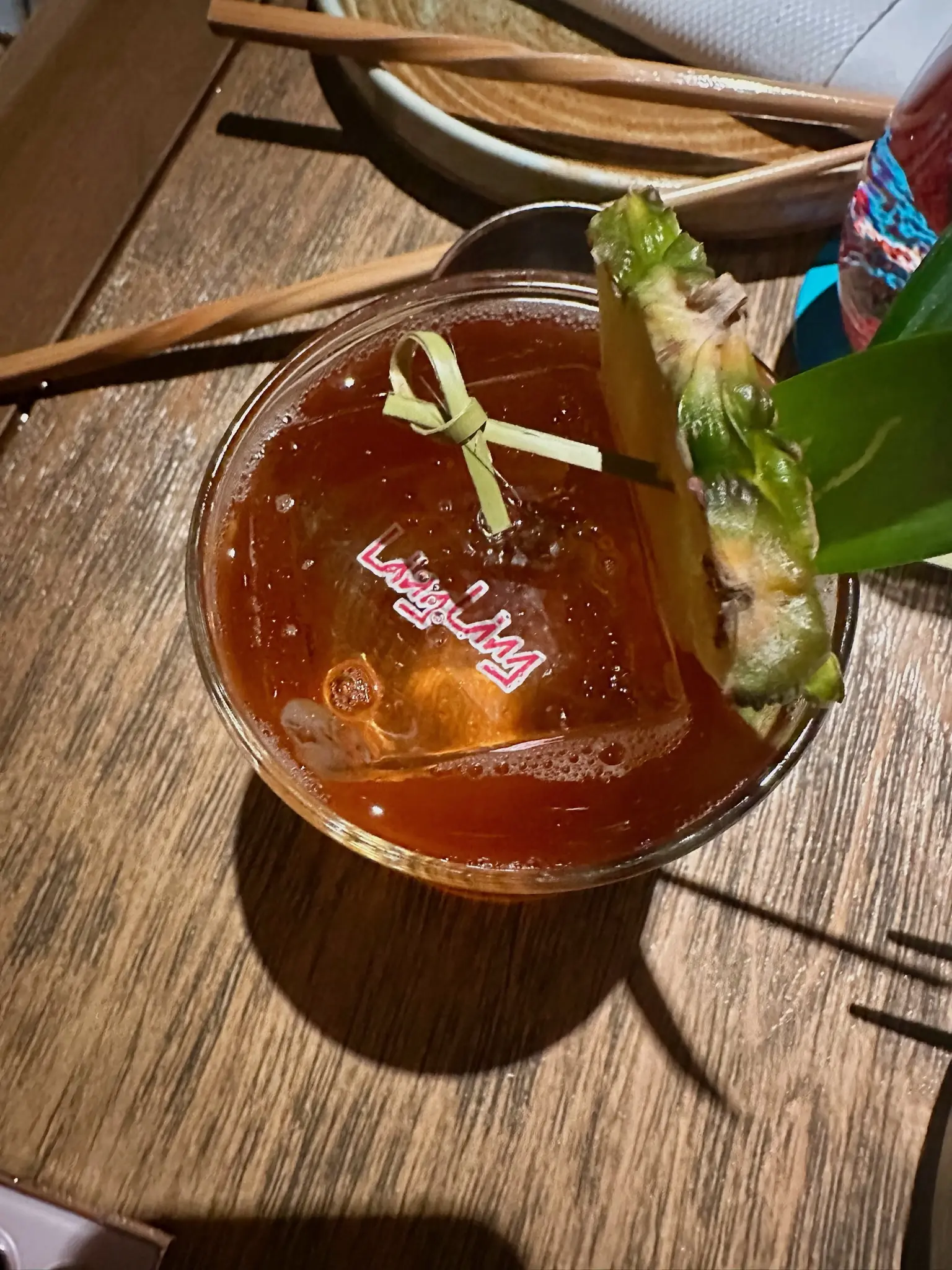 Top down on a redish brown drink with a pineapple slice, agave leaf, and bamboo toothpick in it. A large singular ice cube is in the center of the glass an the words “Ling Ling” appears in the center in pink with a white border.