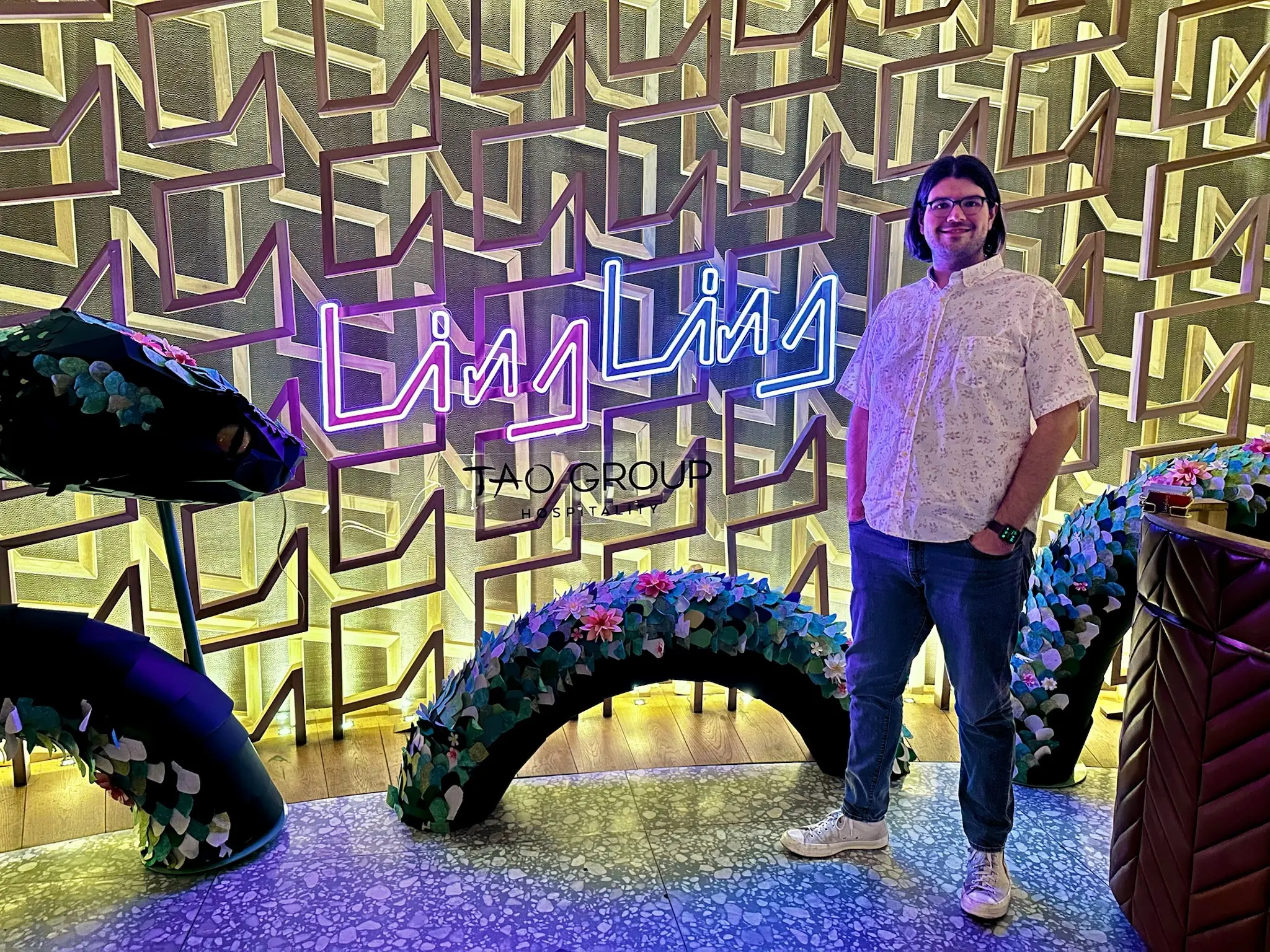 A dimly lit lobby with a patterned, textured wall with a pink and blue neon sign that says “Ling Ling” on the wall. Around the sign is a dragon made out of a topiary that looks like it’s dissapearing into and out of the floor. To the right of the sign is a person (me!) with neck length black hair, blue glasses, and an Apple Watch, wearing a short sleeve white collared shirt, gray jeans, and white Converse high-tops.