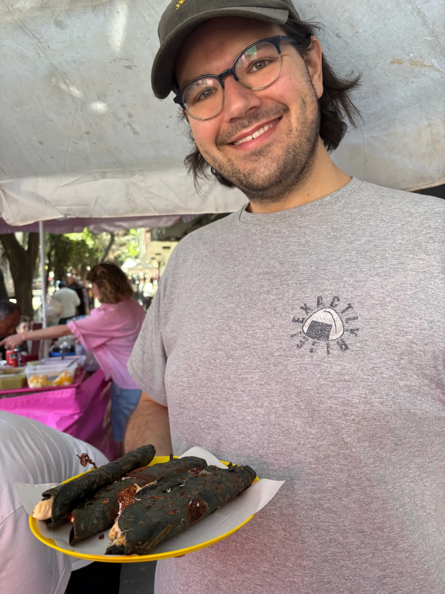 A person (me!) with neck length black hair and blue glasses, wearing a brown courderoy baseball cap with the words “yes, chef” in yellow printed on the front and gray tee-shirt with an onigiri in the upper left with the words “Exactly Rice!” around it, holds a plastic yellow plate with three blue corn quesadillas on it.