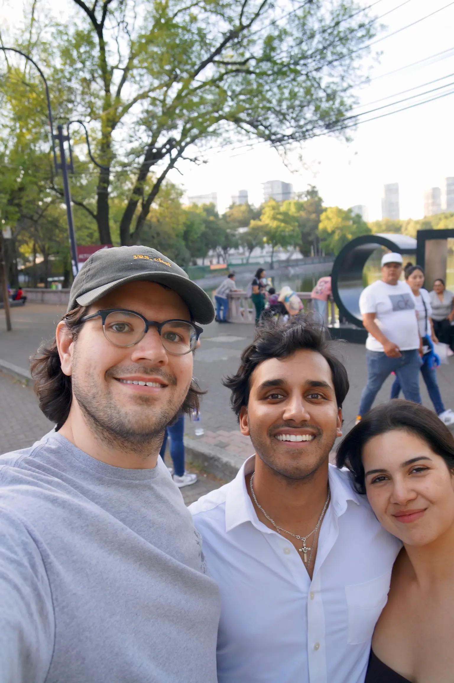 A person (me!) with neck length black hair and blue glasses, wearing a brown courderoy baseball cap with the words “yes, chef” in yellow printed on the front and gray tee-shirt stands next to a man in with shaggy black hair in a long sleeve white collared shirt with a cross necklace on and a woman with long black gair in a maroon dress. Behind them is a park, lake, and cityscape.