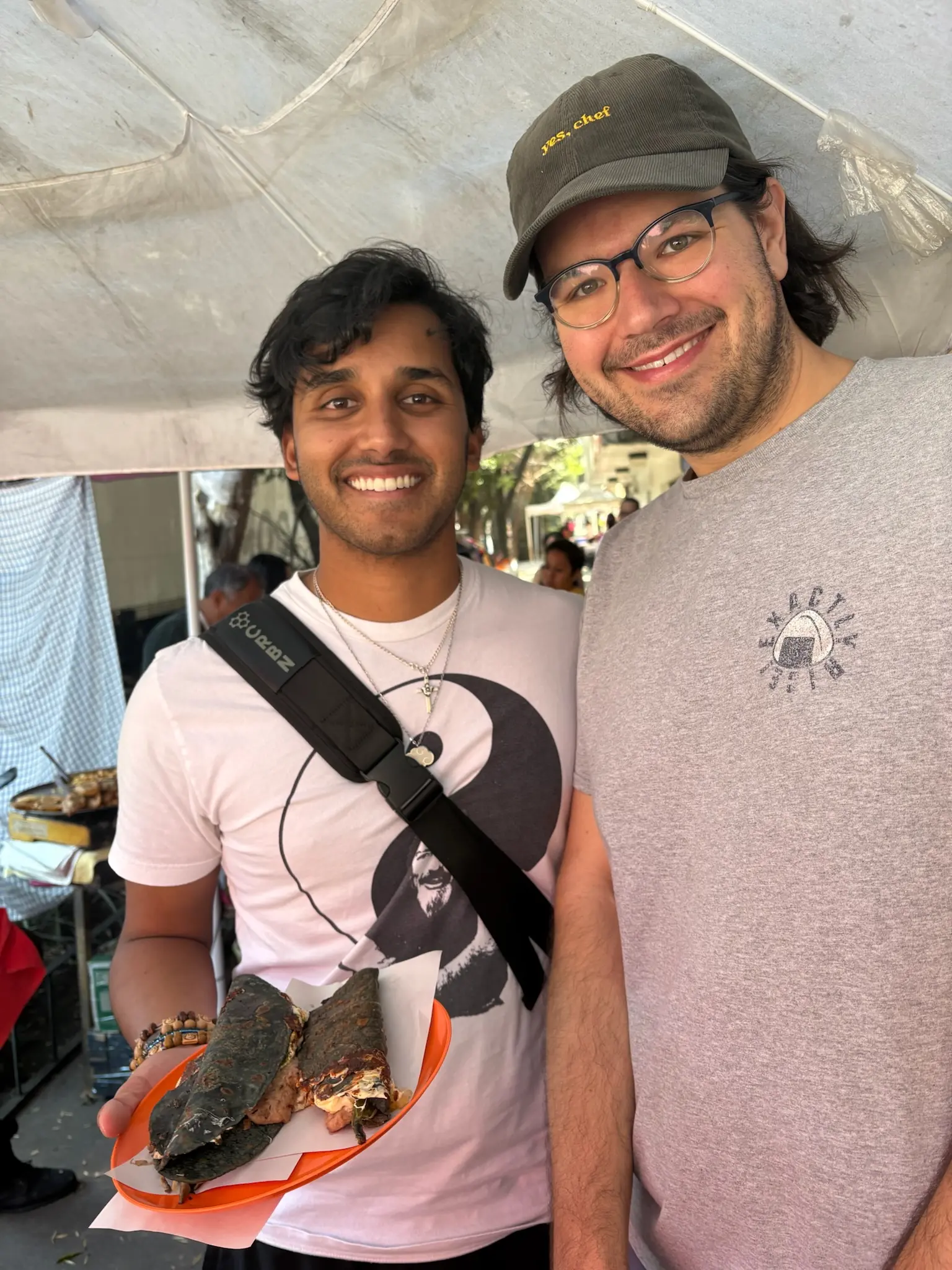 A person with shaggy black hair in a white shirt with a yin-yang on it holds a red plastic plate with two blue corn quesadillas on it. Next to him stands a person (me!) with neck length black hair and blue glasses, wearing a brown courderoy baseball cap with the words “yes, chef” in yellow printed on the front and gray tee-shirt with an onigiri in the upper left with the words “Exactly Rice!” around it.