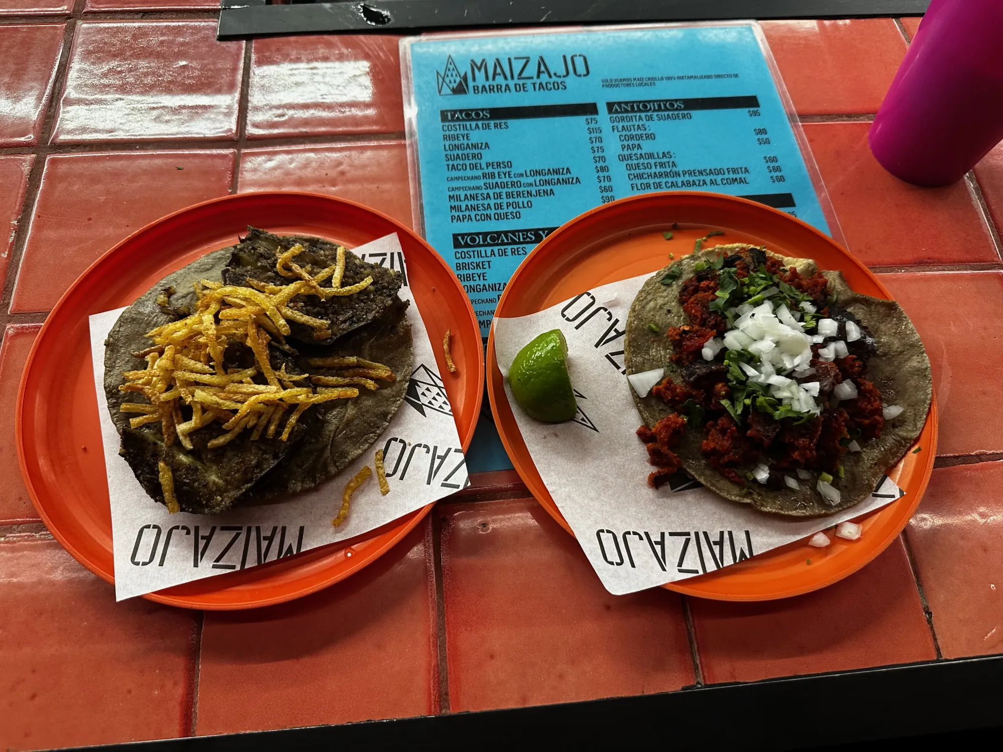On a counter of small, square, red tiles sit two plastic red plates. On top of the plates are a white paper with the word “MAIZAJO” printed on them. On the plate on the left sits a blue corn tortilla topped wih fried eggplant slices and thin slices of crispy fried potatoes. The plate on the right has a lime slice and a taco on a blue corn tortilla with red crumbled longaniza sausage, white onion, and cilantro on top. In the background is a pink cup filled with horchata and a blue menu sits under the plates.