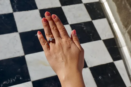 A woman’s hand with pink-painted nails hovers above a black and white checkered floor. On her ring finger is a diamond engagement ring.