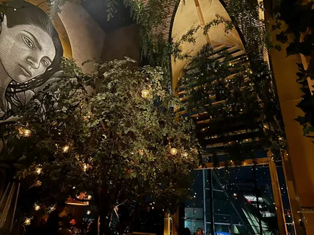 A mural of a woman with long black hair is on a wall with a large lush tree in the center of the room. The window on the right of the room has a large bamboo arch over it with bamboo slits in it.