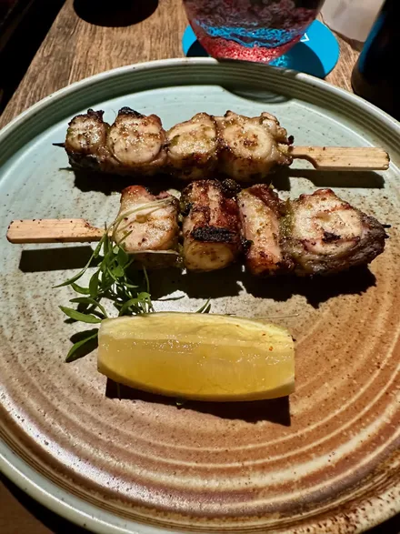 On a round brown ridged plate sit two skewers with four pieces of sliced octopus on each. In front of the skewers is a lemon slice and some sort of green garnish.
