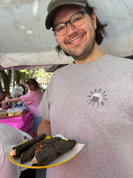 A person (me!) with neck length black hair and blue glasses, wearing a brown courderoy baseball cap with the words “yes, chef” in yellow printed on the front and gray tee-shirt with an onigiri in the upper left with the words “Exactly Rice!” around it, holds a plastic yellow plate with three blue corn quesadillas on it.