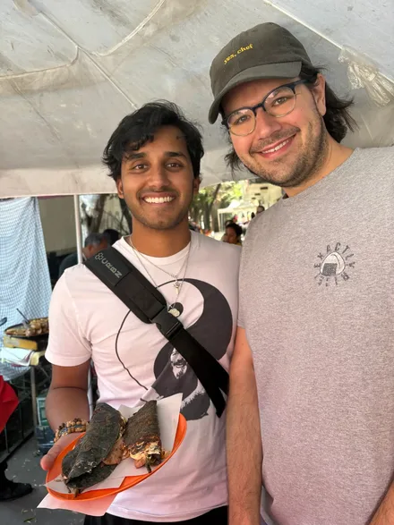 A person with shaggy black hair in a white shirt with a yin-yang on it holds a red plastic plate with two blue corn quesadillas on it. Next to him stands a person (me!) with neck length black hair and blue glasses, wearing a brown courderoy baseball cap with the words “yes, chef” in yellow printed on the front and gray tee-shirt with an onigiri in the upper left with the words “Exactly Rice!” around it.